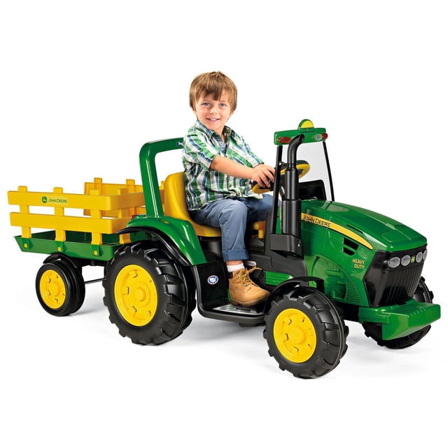Peg Perego John Deere Ride-On Heavy Duty Tractor in green and yellow with a detachable yellow stake-side trailer, large black tires with yellow rims, adjustable yellow seat, windshield, and roll bar, powered by a 12V rechargeable battery for outdoor play.