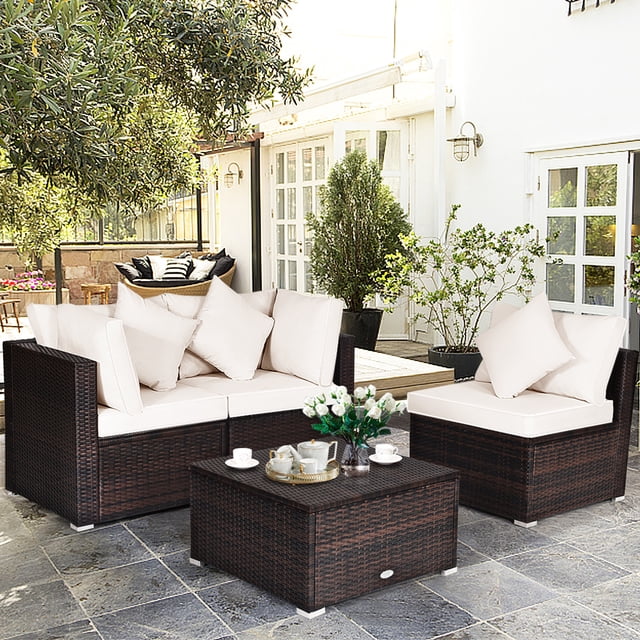 Gymax Ensemble de Conversation pour Patio en Rotin 4 Pcs, Ensemble de Meubles d'Extérieur avec Coussin, Comprenant Deux Canapés d'Angle, un Canapé Sans Accoudoirs et un ottoman. l'Ensemble Est Fabriqué en Rotin PE Brun Résistant aux Intempéries et en Acier, avec des Coussins et des Oreillers Beiges pour le Confort. l'Ottoman Peut Servir de table d'Appoint en Enlevant le Coussin. Exposé dans un Décor de Patio avec de la Verdure et un Plancher de Carreaux.
