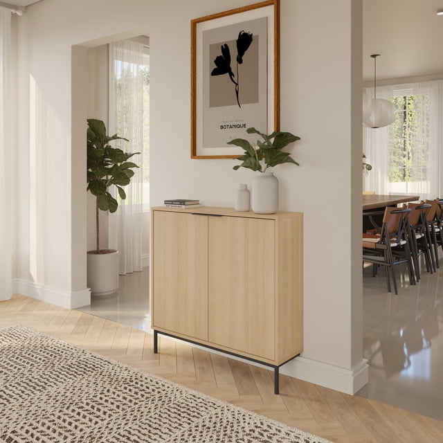 Nexera Slim 2-Door Storage Console in natural blonde oak with black metal hairpin legs, wall-mounted in a bright hallway. The ultra-slim console features two soft-close doors concealing two shelves for tidy storage. Decorated with white vases and a green plant under a framed botanical print. Ideal for narrow spaces and entryways, also suitable for compact storage of everyday essentials.