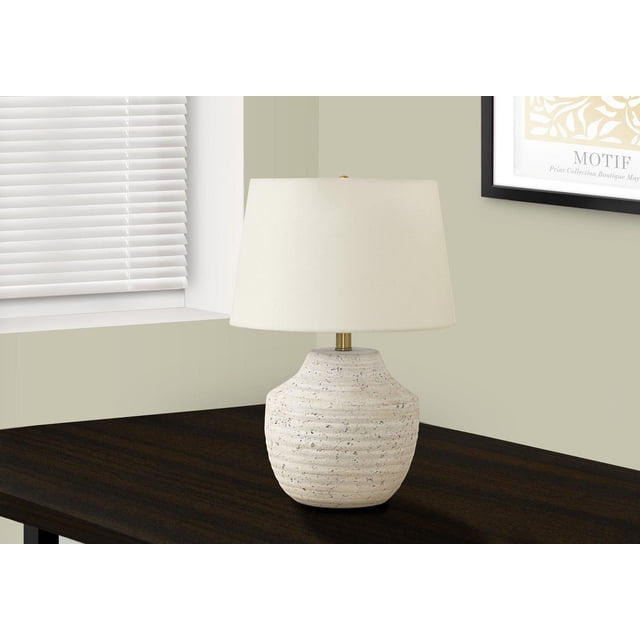 Monarch Specialties Lighting 20"h table lamp with a ridged cream concrete urn-shaped base and textured finish, topped by an ivory linen tapered drum shade and gold finial, placed on a dark wood table near a window with white blinds and framed wall art, ideal for modern living spaces.