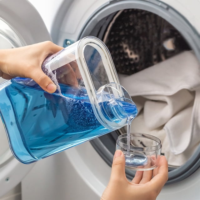 RXIRUCGD Laundry Detergent Dispenser pouring blue liquid detergent into a clear measuring cup. The transparent plastic dispenser, with a wide mouth and sealing lid, is ideal for organizing laundry detergent, softener, and powder. A washing machine with white laundry is visible in the background.