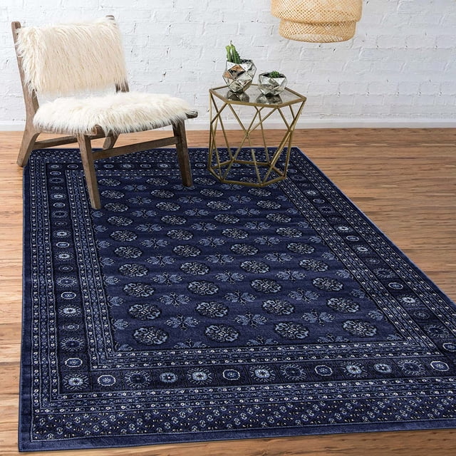 ECARPET Traditional Area Rug with classic oriental Bokhara geometric pattern in deep blue hues, featuring a dense, soft pile suitable for high-traffic areas. The rug lies on a hardwood floor beneath a wooden chair with white faux fur cushion and a gold geometric side table with small plants, set against a white brick wall.