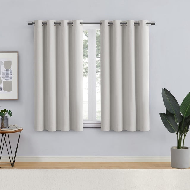 Eclipse Corte 63" Grommet Blackout Window Curtains Panel in off-white blackout fabric with metal grommet top, hanging on a silver rod over a window in a minimalist room with light grey walls, wooden floor, side table with plant, and potted green plant, offering light control and privacy.