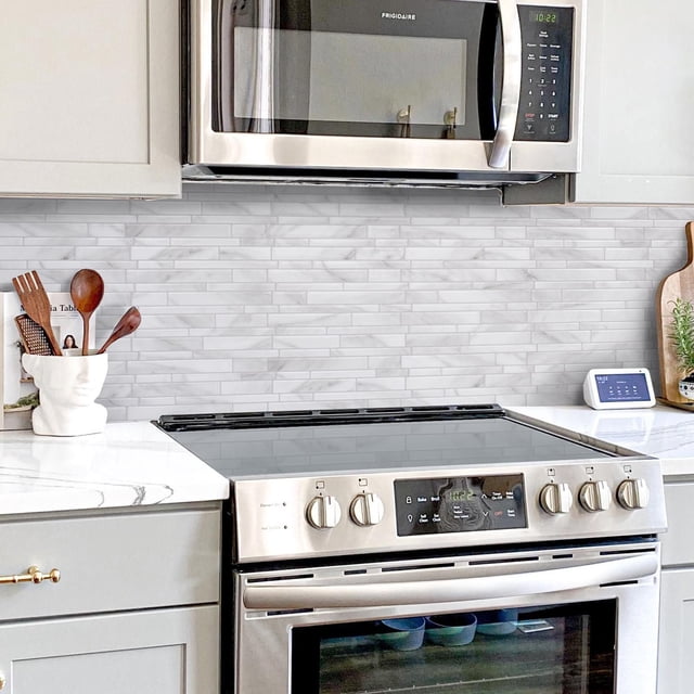 SMART TILES PEEL & STICK BACKSPLASH - MILANO MASSA - 4 Pieces 11.55 x 9.63 installed as a grey marble-patterned backsplash with staggered rectangular tiles behind a stainless steel electric stove and below a Frigidaire microwave, set against white cabinetry and a white countertop with grey veining, featuring wooden utensils in a white face-shaped holder and a smart home display on the counter, suitable for kitchen and bathroom wall applications.