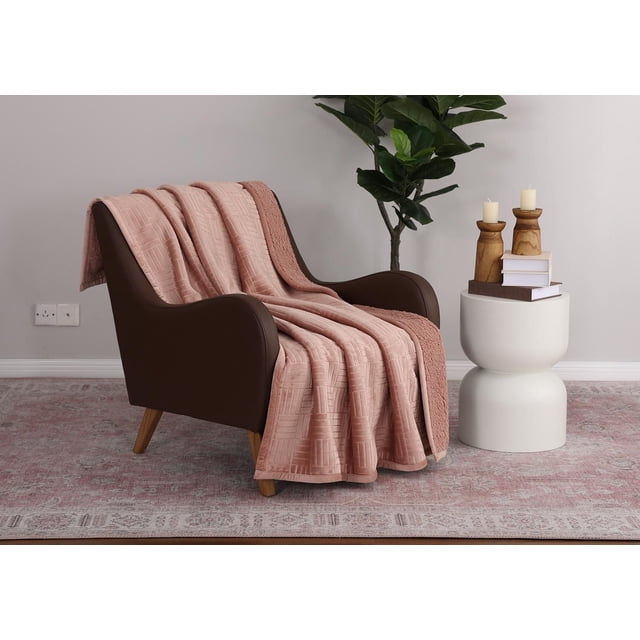 Oversized Plush Throw in muted pink with embossed velvet plush texture and faux shearling reverse, draped over a dark brown armchair with wooden legs. The 50 in. x 70 in. throw is displayed in a living room setting with a white side table holding books and wooden candle holders, a green plant, and a patterned pink and beige rug. It is often used for added warmth and comfort during relaxation.