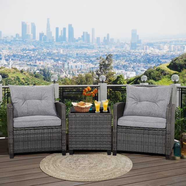 Gymax 3PCS Patio Rattan Sofa Set Outdoor Wicker Conversation Set with a mix grey PE wicker design and grey cushions. Includes two single chairs with ergonomic curved armrests and backrests, and a compact coffee table with a black tempered glass tabletop. The set is displayed on a wooden patio with a cityscape view in the background.