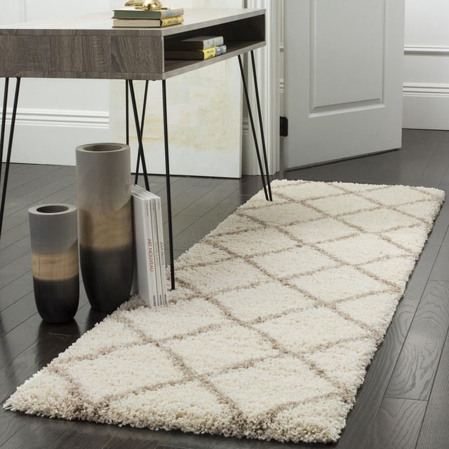 Safavieh Hudson Amias Geometric Shag Area Rug in ivory with beige onion-shaped tile patterns on a thick plush shag pile, placed on dark hardwood flooring next to a contemporary wood console table with black metal hairpin legs, flanked by two gradient ceramic vases and books, suitable for high-traffic urban or modern living spaces.