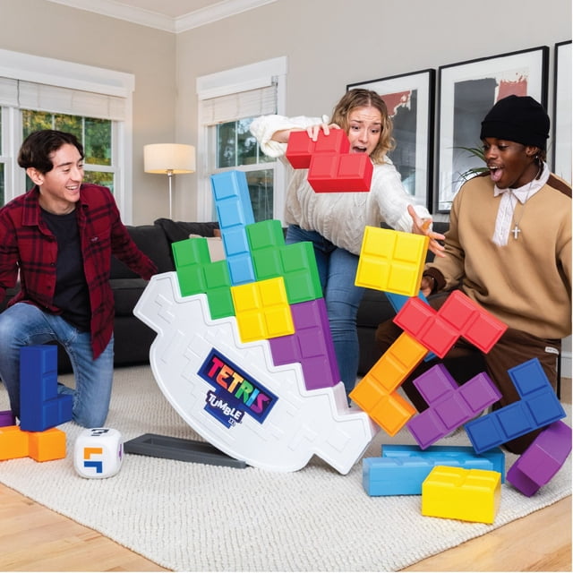 Tetris XL Tumble Game by EastPoint Sports displayed in a living room with three players actively stacking oversized, colorful Tetrimino blocks on a white tilting base labeled 'Tetris Tumble XL.' The game includes blocks in red, blue, green, yellow, orange, and purple, and a large die with Tetris symbols is visible on the floor. The tilting base sways as the tower grows, adding a dynamic challenge. Suitable for solo or head-to-head play, the game can stack up to 6 feet high.