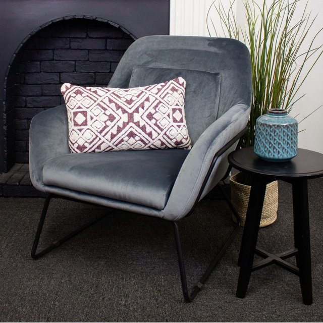Dolce Luxury Cushion Cover (no insert included) on a grey velvet armchair with black metal legs, featuring a 12x20 inch rectangular cushion cover in white and burgundy geometric pattern, suitable as a decorative throw pillow cover for home décor, shown in a modern room with a black brick fireplace, black round side table with a blue textured ceramic vase, and tall green plants in a woven basket.