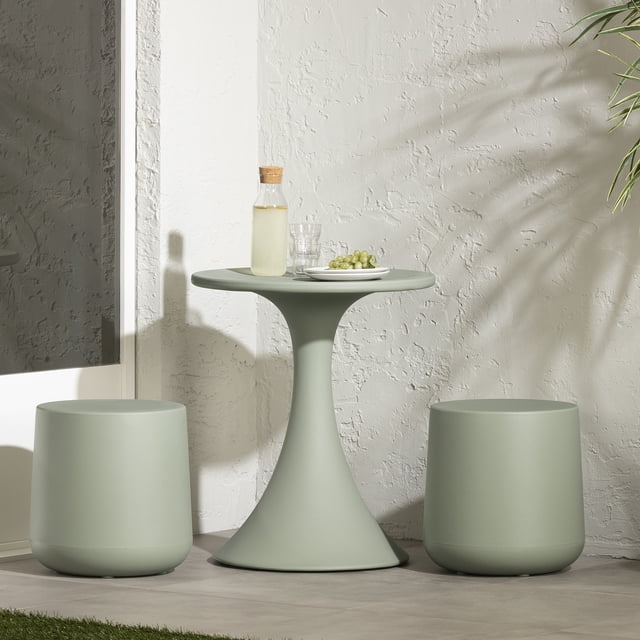 South Shore Dalya Table and Stools Bistro Set in sage green with a compact round pedestal table and two rounded stools made of durable polyethylene, shown with a glass carafe, glass, and plate of grapes on the table against a textured white wall in a minimalist patio setting, suitable for small outdoor spaces.