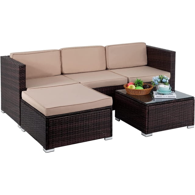 FDW 5-Piece Outdoor Patio Set featuring a sectional rattan wicker sofa with khaki cushions, a matching ottoman, and a tempered glass-top table. The set is designed for outdoor use in gardens, porches, balconies, or poolside areas. The steel frame provides stability, and the PE rattan wicker ensures durability in all weather conditions. The table is styled with a basket of fruit, a cup, and books for decorative purposes.