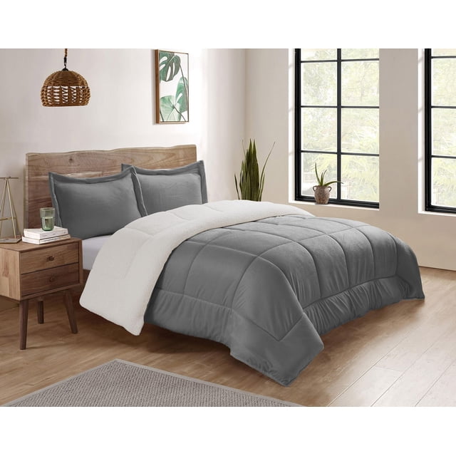 Reversible Mink & Berber Plush Sherpa Comforter Set in grey on a bed with a natural wood headboard, showing the plush grey side and folded back white Sherpa fleece side, with matching grey pillow shams, in a bright bedroom featuring wooden flooring, a wooden nightstand with books and a glass, and large black-framed windows overlooking greenery, suitable for cozy warmth and comfort.