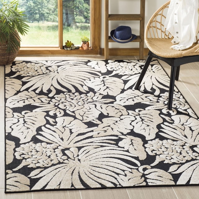 SAFAVIEH Monroe Zavia Floral Area Rug with a black background and beige floral patterns, machine-loomed from durable synthetic yarns for indoor and outdoor use, shown on light wood tile flooring with a rattan chair and wooden shelf, suitable for high-traffic areas and decorative accent use.