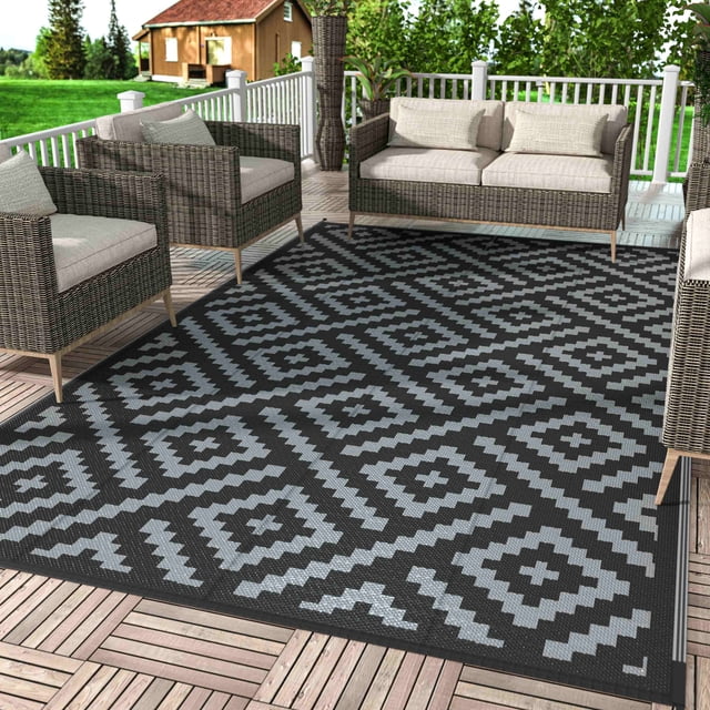 DEORAB Outdoor Rug Reversible 9x12' Patio Rug in black and grey, featuring a geometric lines pattern. This waterproof, UV-resistant outdoor area rug is displayed on a wooden patio surrounded by wicker furniture. Lightweight and foldable, it is easy to clean and ideal for patios, camping, RVs, decks, porches, balconies, backyards, and picnics.