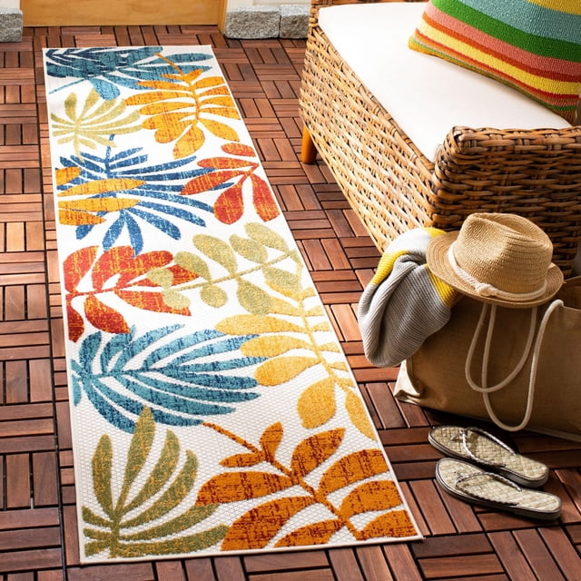 SAFAVIEH Cabana Elouise Floral Indoor/Outdoor Area Rug with a white background and colorful tropical leaf patterns in blue, orange, yellow, and green. This durable synthetic rug is placed on a wooden patio floor next to a wicker sofa with a white cushion and striped pillow, accompanied by a straw hat, tote bag, blanket, and sandals, ideal for outdoor living spaces.