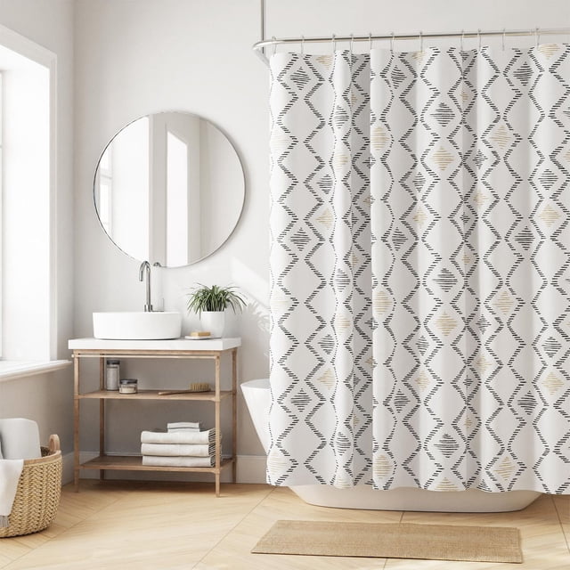 Hometrends Recycled Fabric Decorative Shower Curtain with black and beige geometric diamond patterns on white recycled fabric, hanging from a curved rod in a bright bathroom with a round mirror, wooden vanity with white vessel sink, woven basket, and beige bath mat.