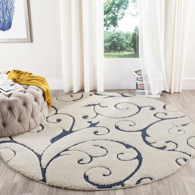 SAFAVIEH Florida Douglas Floral Vines Shag Area Rug, round with plush high-low pile in crème and dark blue damask vine pattern, on light wood flooring near a beige tufted ottoman and sheer white curtains with a view of green trees, ideal for adding softness and texture to living areas.