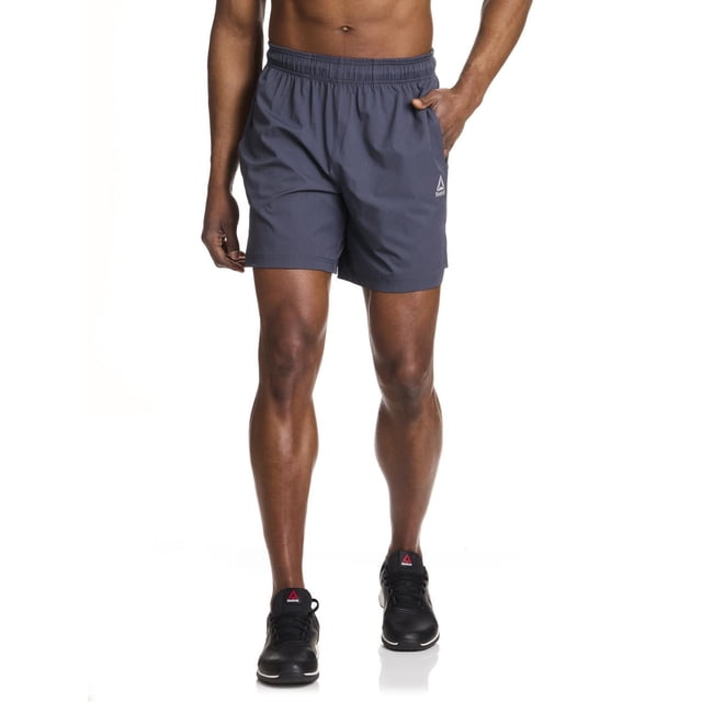 Reebok Men's Pace Breaker Training Short in grey with a 7-inch inseam, shown front-facing on a male model. Made from breathable 89% polyester and 11% spandex stretch fabric, these performance shorts feature an elastic waistband, bonded zip side pocket, and Reebok Delta logo. Ideal for high-intensity training and casual wear, they provide flexibility and secure storage.