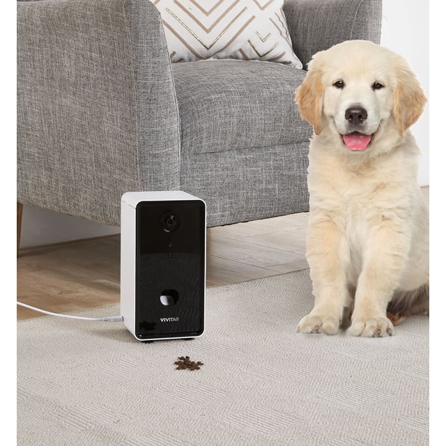 Distributeur Intelligent pour Animaux par Vivitar sur un Tapis beige Pâle à Côté d'Un Fauteuil Rembourré Gris avec un Oreiller à Motifs, avec un Panneau avant Noir avec un Objectif de Caméra 4K et une Fente de Distribution de Collations, avec des Gâteries pour Chiens sur le Sol et un Chiot golden retriever Assis à Proximité dans un Salon.