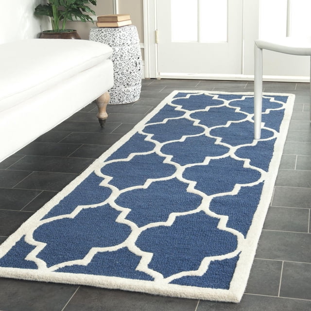 Safavieh Cambridge Kevin Geometric Area Rug with blue hand-tufted wool and white quatrefoil pattern, placed on dark tile floor in a bright room featuring a white sofa and decorative ceramic stool with books, suitable for modern home or office accent.