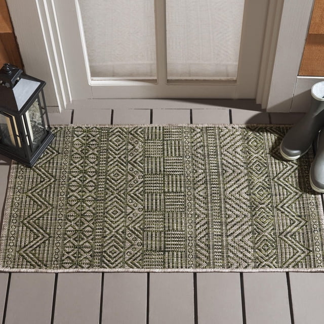 SAFAVIEH Courtyard Altan Chevron Indoor/Outdoor Area Rug with a rectangular shape featuring green and beige chevron and geometric patterns woven in durable polypropylene sisal weave, placed on a gray wooden porch floor in front of a glass door with a black lantern and green rubber boots, suitable for indoor and outdoor use.