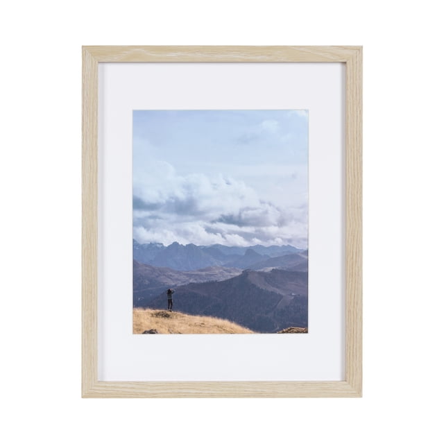 hometrends Gallery Natural Picture Frame with light natural wood finish and white mat, displaying a vertical photo of a person standing on a grassy hill overlooking mountain ranges under a cloudy sky, suitable for tabletop or wall display with included easel and hangers, often used to showcase memorable moments or as a personalized gift.