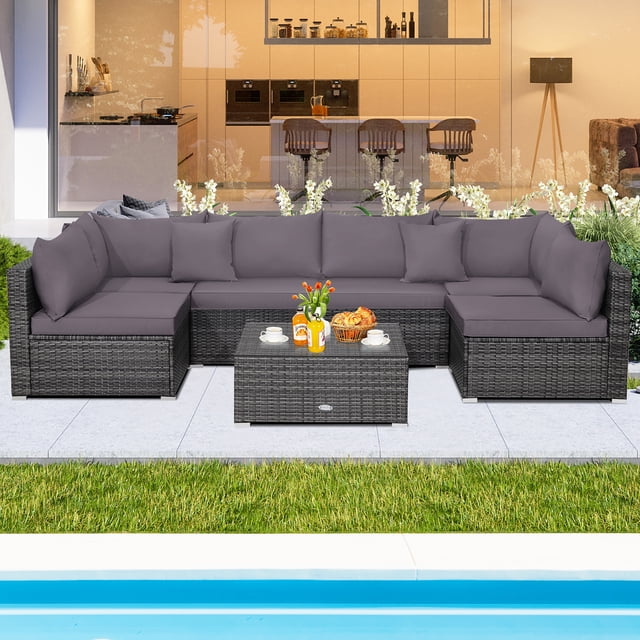 Costway 7PCS Patio Rattan Furniture Set Sectional Sofa Cushioned Garden Grey displayed in an outdoor setting. The set includes a mix grey PE rattan sectional sofa with grey cushions, a tempered glass coffee table, and two throw pillows. The modular design features two corner sofas, four armless sofas, and a table, allowing flexible arrangements. The furniture is weather-resistant with a rustproof steel frame and anti-rust aluminum feet, suitable for patios, gardens, or poolside areas. The cushions are thickly padded for comfort, with removable covers for easy cleaning.