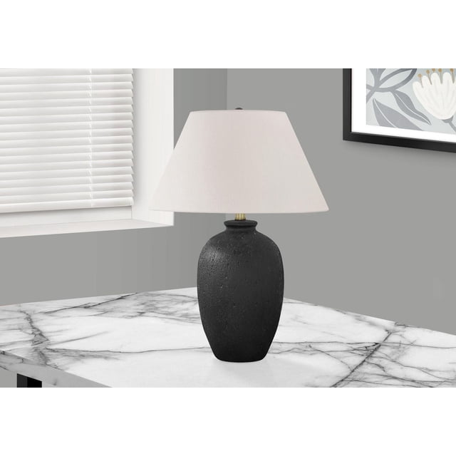 Monarch Specialties Lighting 24"h table lamp with a textured black ceramic vase-shaped base, ivory linen empire shade, and round black finial, placed on a white marble table in a modern gray room with a window and framed artwork, suitable for bedroom or office use.