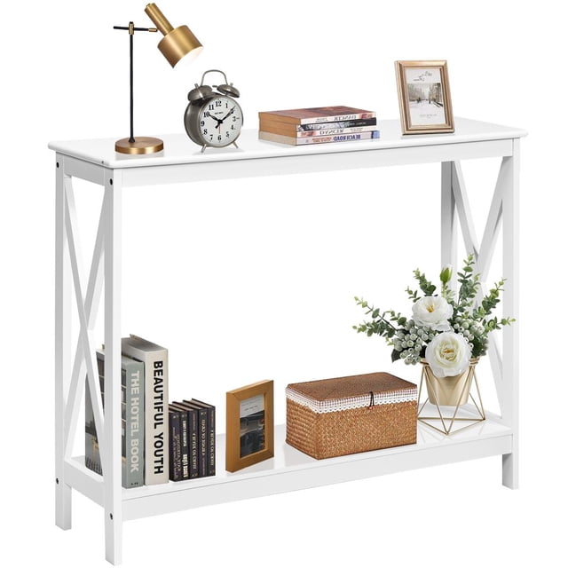 Giantex console table in white with a narrow design, X-shaped side frames, and two tiers for storage. The tabletop displays decorative items such as a lamp, clock, books, and a framed photo, while the lower shelf holds books, a wicker box, and a flower arrangement. This modern accent table is ideal for entryways, living rooms, or offices.