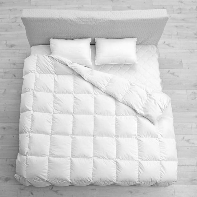 Ambassador Microfiber Duvet - 233 Thread Count on a bed with a light grey quilted headboard and wooden floor. The white duvet features square baffle box stitching with one corner folded back, showing its soft gel microfiber fill and breathable cotton shell. Two white pillows rest at the head, emphasizing its hypoallergenic and even heat distribution properties for comfortable sleep.