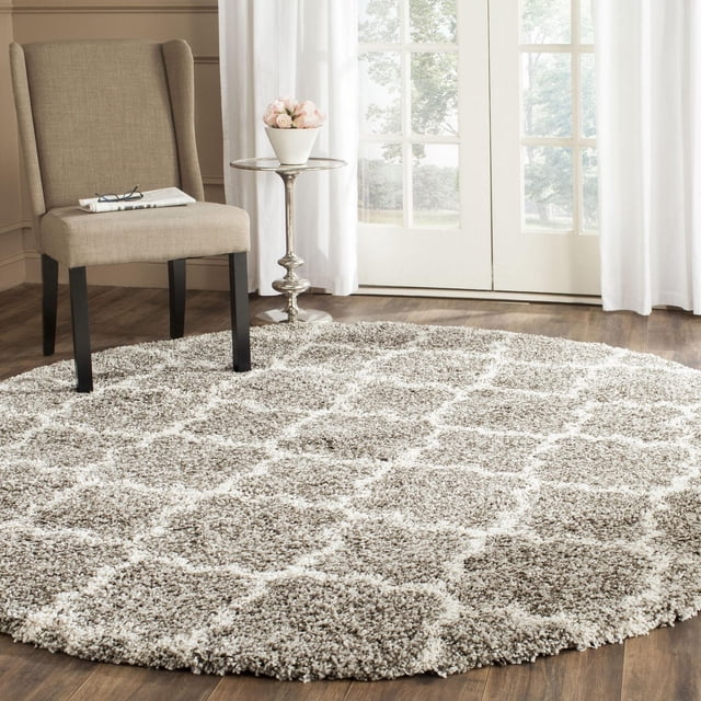Safavieh Hudson Jaye Tapis Shag Géométrique pour Aire, Rond avec une Épaisse Peluche Shag en Motifs de Carreaux Ivoire et taupe, Posé sur Plancher de Bois Franc à Côté d'Une Chaise Rembourrée beige et d'Une table d'Appoint Argentée avec un vase Blanc de Fleurs Roses, dans une Pièce Lumineuse avec des Rideaux Blancs et des Portes en Verre. Ce Tapis d'Accent Ajoute de la texture au décor Moderne et Convient aux Espaces de Vie Très Passants.