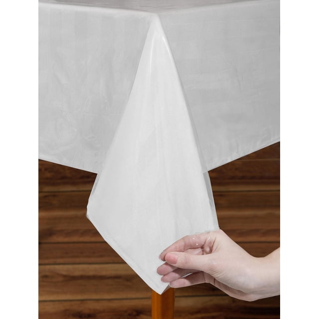 Mainstays Protège-Nappe Robuste, Imperméable, Protecteur en Plastique Transparent Montré Drapé sur un Coin de table en Bois avec une Main Soulevant le Bord pour Révéler Sa texture Imperméable, Conçu pour Protéger le Linge de table et les tables contre les Déversements et les Dommages, Convient à Diverses Formes et Tailles de table.
