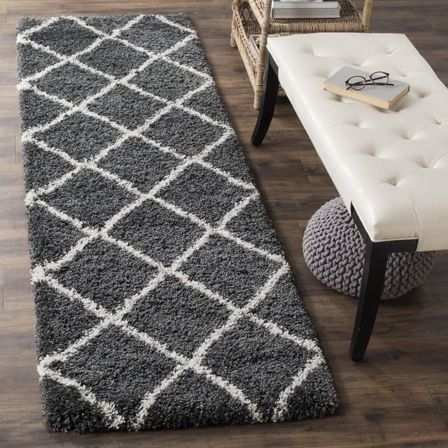 Safavieh Hudson Amias Geometric Shag Area Rug runner featuring a plush dark grey shag pile with white diamond tile patterns, placed on wood flooring beside a white tufted bench and wicker table, ideal for adding texture and durability to modern living spaces.
