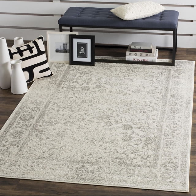 SAFAVIEH Adirondack Wyatt Traditional Area Rug in light beige with subtle antiqued floral and border patterns, placed on dark hardwood flooring. Styled with a navy tufted bench, cream pillow, black and white framed art, and white ceramic vases, suitable as a durable accent rug for rustic or modern home interiors.