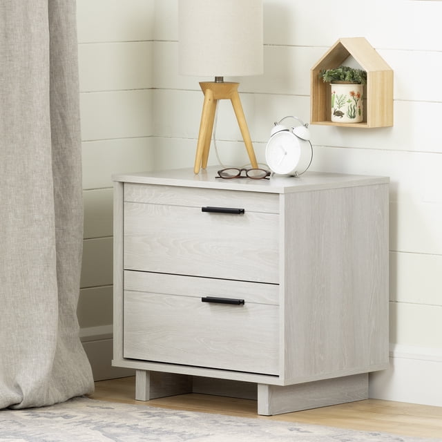 South Shore Fynn 2-Drawer Nightstand in winter oak with two spacious drawers featuring black metal handles and smooth metal slides, placed in a child's bedroom corner with a beige curtain, wooden tripod lamp, white alarm clock, and a wooden house-shaped wall shelf holding a plant pot, suitable for storing bedtime essentials.