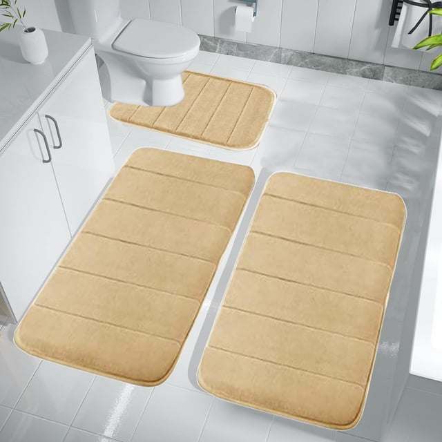Dvkptbk Memory Foam Bath Mat Set with three khaki-colored rugs, including a toilet mat, bath mat, and floor mat. Made with velvet fleece and memory foam for softness, water absorption, and non-slip backing. Machine washable and designed for comfort. Displayed in a modern bathroom with white tiles and fixtures.