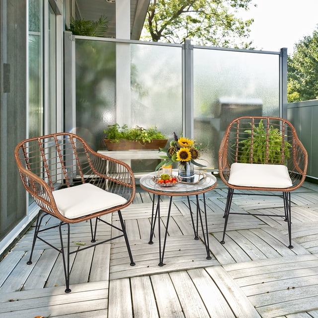 Costway 3PCS Patio Rattan Bistro Set with two armchairs featuring beige cushions and a round tempered glass coffee table. Perfect for compact outdoor spaces like patios, gardens, balconies, or poolside. The set is crafted with durable rattan and iron legs, displayed on a wooden deck with greenery and sunlight.