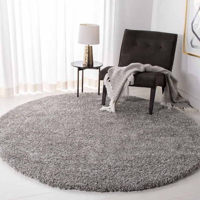 Safavieh California Solid Plush Shag Area Rug, round with a dense 4.5kg/m² polypropylene pile in soft grey, placed on a light floor beside a dark upholstered chair with a patterned throw and a small round side table with a gold textured lamp, suitable as a plush accent rug for home interiors.