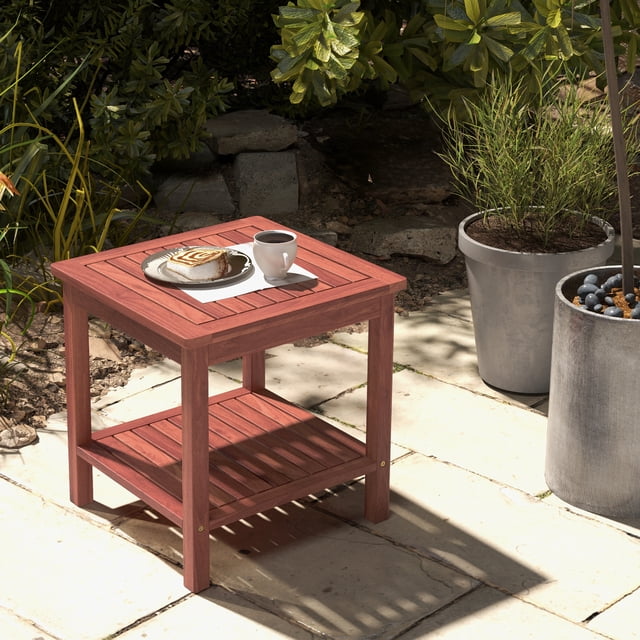Patiojoy 2pcs Patio Side Table Double-Tier Acacia Wood End Table with a slatted tabletop and bottom shelf, shown in a natural wood finish. The table is placed outdoors on a stone patio surrounded by greenery and potted plants. A cup of coffee and a plate with a pastry are on the tabletop, highlighting its functionality for outdoor relaxation.