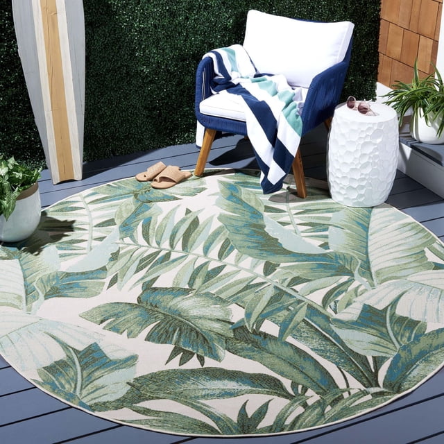 SAFAVIEH Barbados Igor Tapis Botanique Intérieur/extérieur de Forme Ronde avec Motif de Feuilles Tropicales Vertes et Bleues sur un Fond Clair, Exposé sur une Terrasse Extérieure Grise avec un Fauteuil Bleu Marine avec des Coussins Blancs et un Jeté à Rayures, à Côté d'Une table d'Appoint Texturée Blanche avec des Lunettes de Soleil et des Plantes en Pot, Idéal pour Ajouter des vibes Côtières aux Espaces de patio.