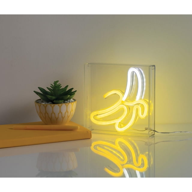 Merkury Innovations 6" Banana LED Neon Box with bright yellow and white LED lights shaped like a peeled banana inside a clear polycarbonate 6x6 inch box, USB-powered and perfect for adding character to desks or shelves, shown on a white surface next to a green succulent in a white and yellow pot and a yellow notebook with pen.