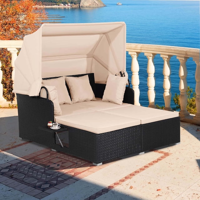 Costway Patio Rattan Daybed Lounge with retractable beige canopy, black hand-woven PE wicker frame, and beige cushions. Features two retractable side tables and includes four throw pillows. Designed for outdoor use with a rustproof steel frame and a weight capacity of 800 lbs. Suitable for patios, gardens, and poolside settings.