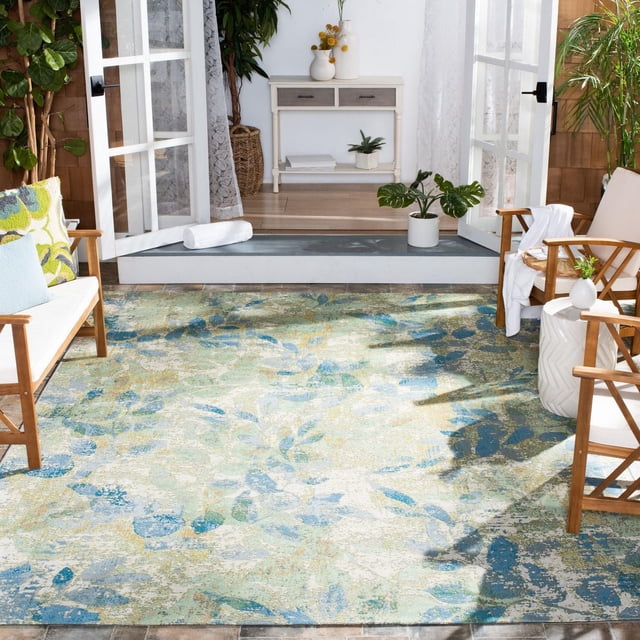 SAFAVIEH Barbados Gresham Botanical Indoor/Outdoor Area Rug featuring a blue and green botanical leaf pattern on a textured surface, placed on a patio between wooden chairs with white cushions and light blue pillows. The scene includes open glass doors, potted plants, and a white console table indoors, ideal for durable outdoor decor with coastal vibes and suitable for enhancing patio living spaces.