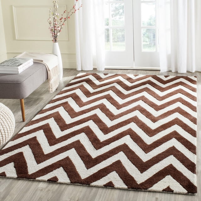 Safavieh Cambridge Kaitlyn Zig Zag Stripes Area Rug with brown and white zigzag pattern and hand-tufted wool texture, displayed on light wood flooring in a bright room with a gray bench, beige throw, white vase with flowers, and sheer white curtains, ideal for adding modern American style to home decor.