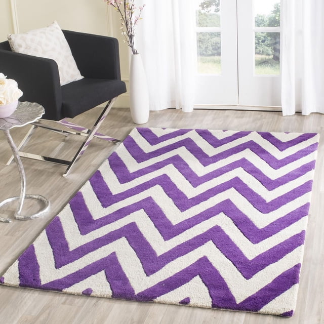 Safavieh Cambridge Kaitlyn Zig Zag Stripes Area Rug with purple and white zigzag stripes, hand-tufted wool plush cut pile, displayed on light wood flooring in a bright room with a black armchair, white patterned pillow, and tall white vase with flowers near glass doors with white curtains, ideal for modern home accent.