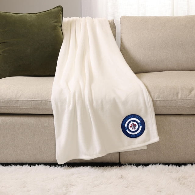NHL, NBA, MLB White Sports Throw Blanket, 50" x 60", plush 100% polyester, draped over a beige sofa with a green pillow, featuring a Winnipeg Jets logo patch in blue and red near the bottom corner, ideal for cozy indoor warmth and versatile use as a throw blanket.