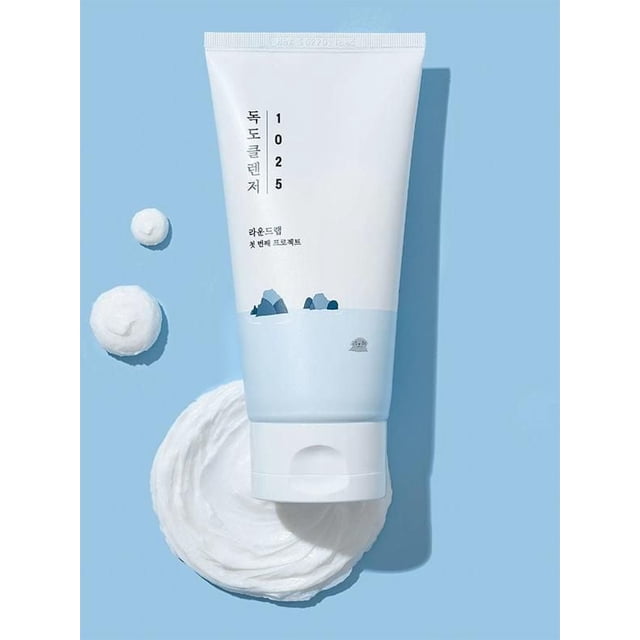 Round Lab 1025 Dokdo Cleanser in a white and light blue tube with Korean text and mountain graphics, placed on a light blue background with thick white foamy cleanser swirled beside it, highlighting its hydrating, gentle formula ideal for sensitive skin and effective makeup removal.