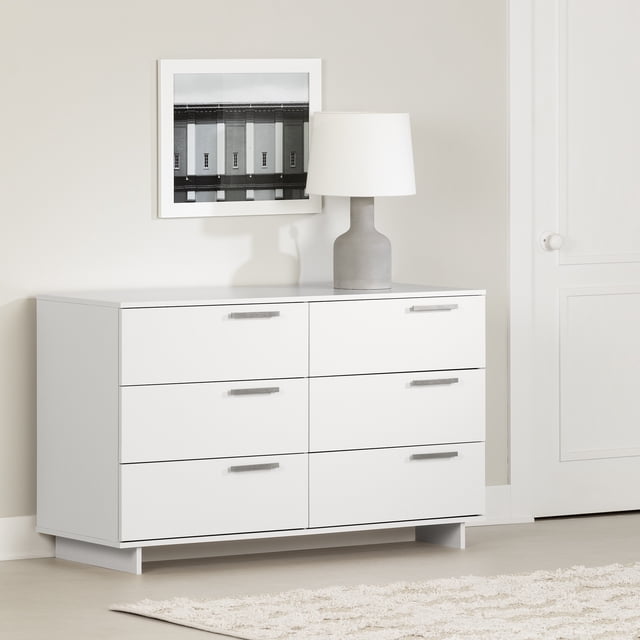 South Shore Cavalleri 6-Drawer Dresser for Adult in pure white featuring six spacious drawers with off-center metal handles and metal slides for durability. The modern dresser has slim legs creating a floating effect and a flat top surface suitable for bedroom storage or display, set against a beige wall with a framed photo and grey lamp.