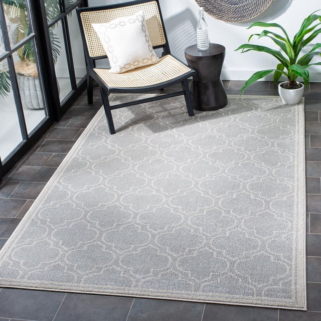 Safavieh Amherst Wilson Geometric Area Rug in light grey with white trellis pattern on dark grey tile floor, styled with a black-framed woven chair with a white chain-link patterned cushion, a black side table with a glass bottle, and a green potted plant, suitable for high-traffic indoor-outdoor areas.