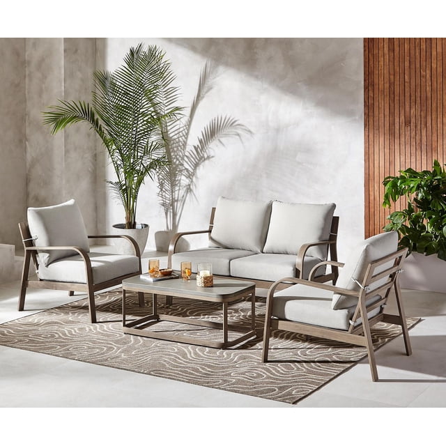 Better Homes & Gardens Prado 4-Piece Steel Patio Outdoor Conversation Set, Brown, with a loveseat and two club chairs featuring UV- and fade-resistant light grey cushions on rust-resistant, powder-coated steel frames with faux wood finish, arranged around a rectangular steel coffee table with hand-painted concrete finish on a patterned outdoor rug in a sunlit patio with potted plants and textured walls, ideal for durable and comfortable outdoor seating.